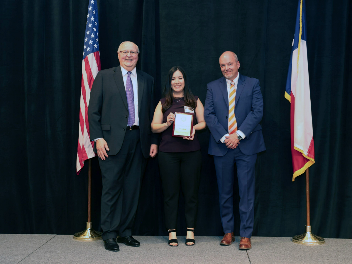 Three people in business attire stand on a stage before a black curtain; the person in the center holds an award plaque, with U.S. and Texas flags on either side of the group.
